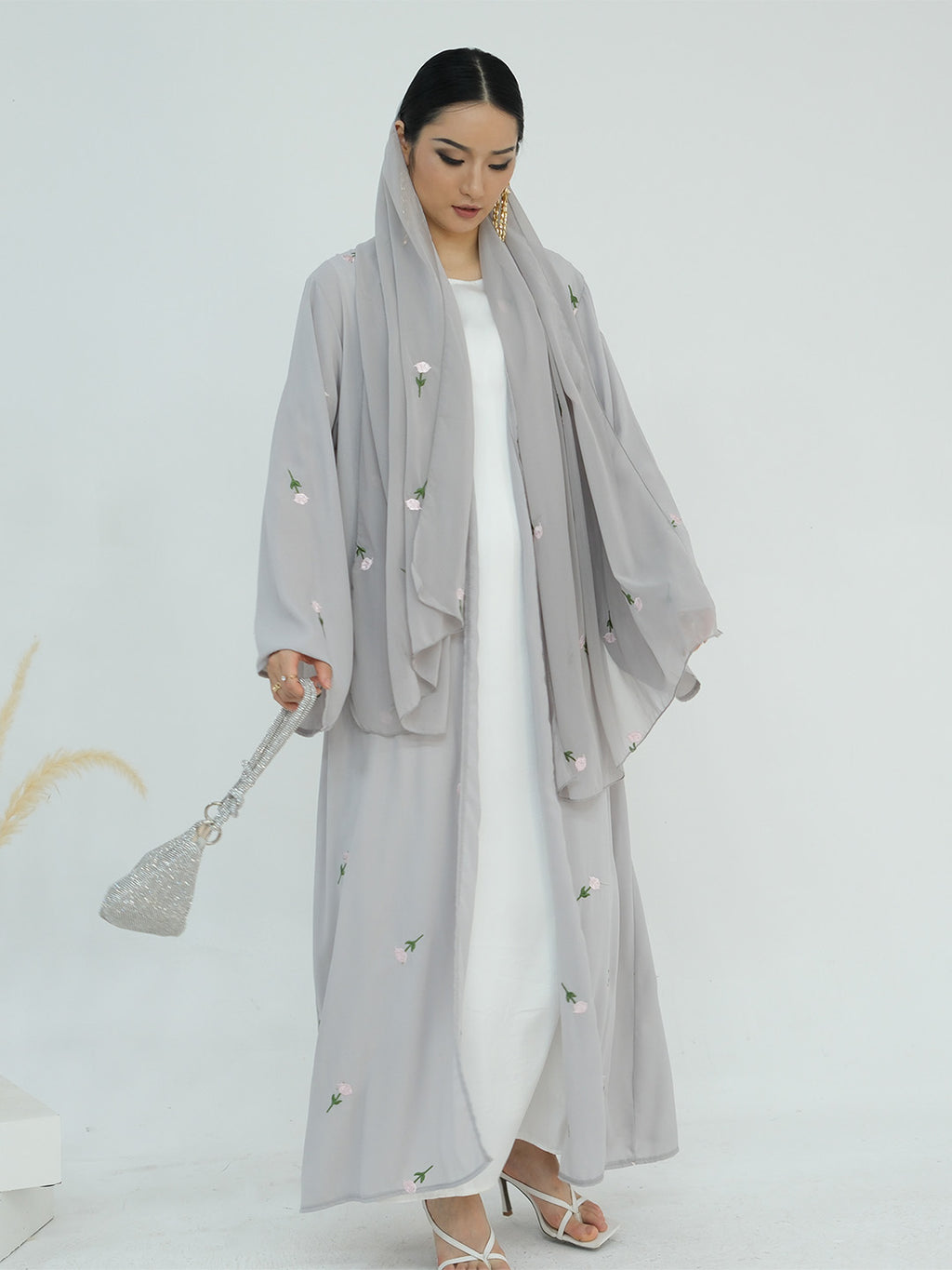 A woman modeling a light gray modest outfit consisting of a long open-front abaya with embroidered details, a matching headscarf, and a white inner dress, holding a sparkly silver handbag against a plain white studio background.
