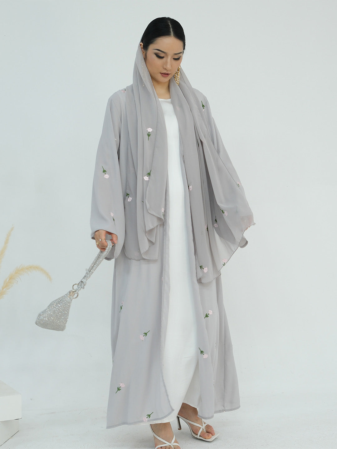 A woman modeling a light gray modest outfit consisting of a long open-front abaya with embroidered details, a matching headscarf, and a white inner dress, holding a sparkly silver handbag against a plain white studio background.