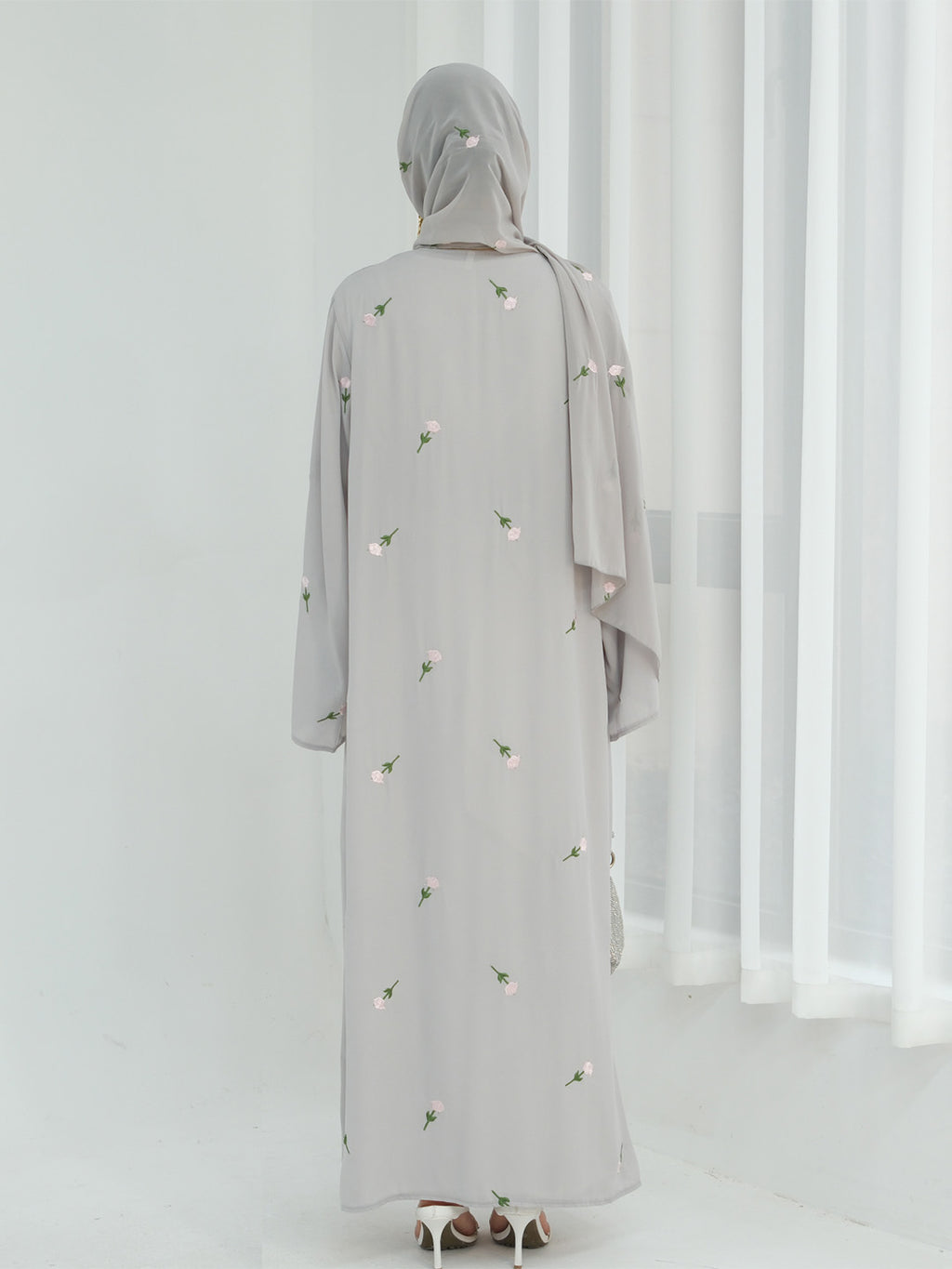 A woman viewed from behind wearing a light gray abaya with small white floral embroidery and a matching gray hijab, standing in a bright white room with sheer curtains.
