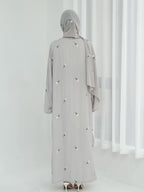 A woman viewed from behind wearing a light gray abaya with small white floral embroidery and a matching gray hijab, standing in a bright white room with sheer curtains.