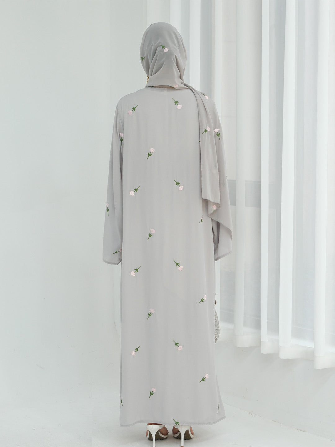 A woman viewed from behind wearing a light gray abaya with small white floral embroidery and a matching gray hijab, standing in a bright white room with sheer curtains.