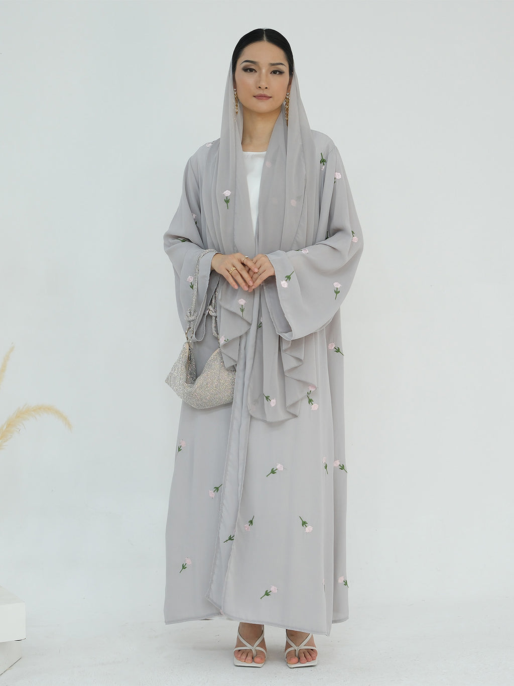 A woman modeling a light gray modest outfit featuring a long-sleeved embroidered abaya with a matching headscarf and a small woven handbag, set against a plain white studio background.