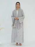 A woman modeling a light gray modest outfit featuring a long-sleeved embroidered abaya with a matching headscarf and a small woven handbag, set against a plain white studio background.