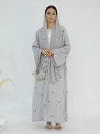 A woman modeling a light gray modest outfit featuring a long-sleeved embroidered abaya with a matching headscarf and a small woven handbag, set against a plain white studio background.