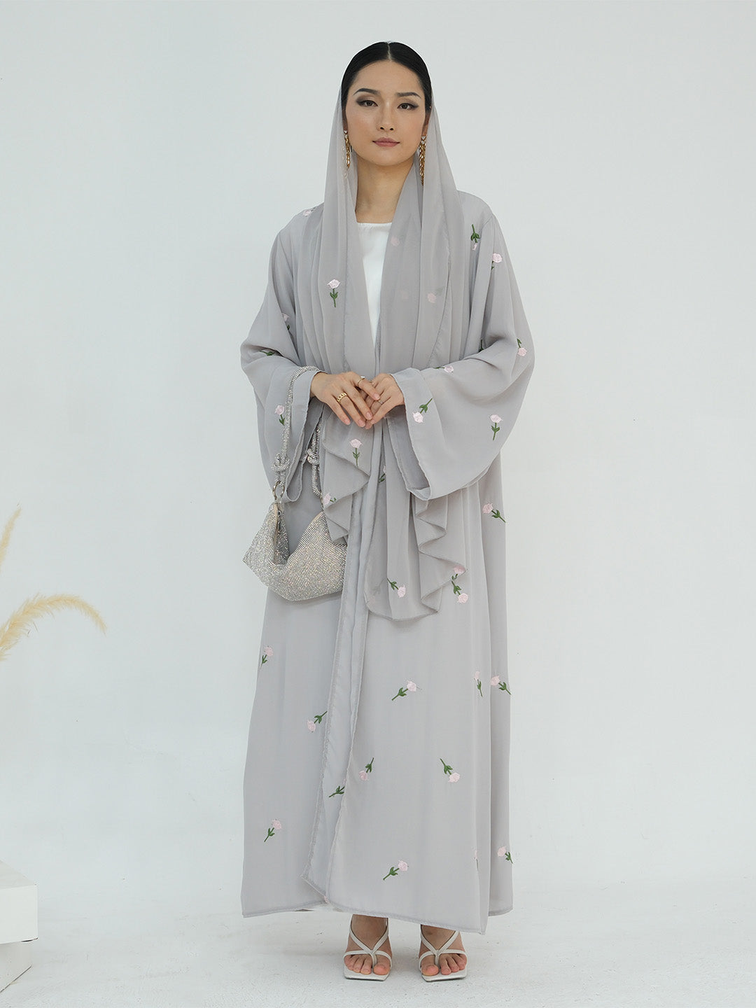 A woman modeling a light gray modest outfit featuring a long-sleeved embroidered abaya with a matching headscarf and a small woven handbag, set against a plain white studio background.