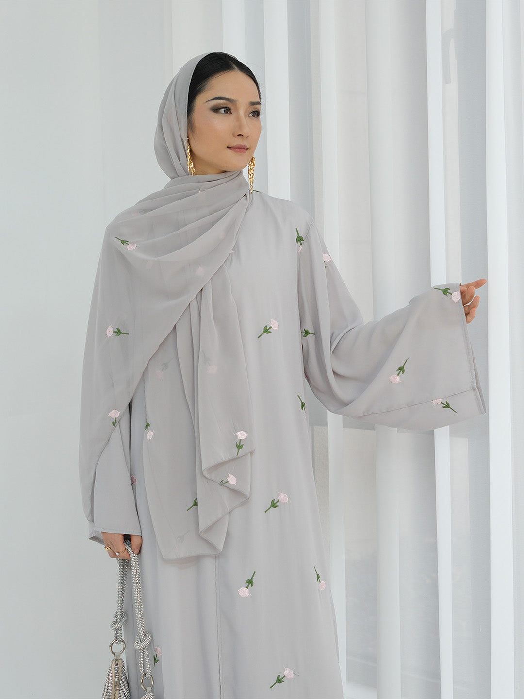 A woman wearing a light gray modest outfit with a matching draped hijab, featuring small embroidered white flowers and green leaves. She is standing in front of white vertical drapes, holding a silver chain handbag.