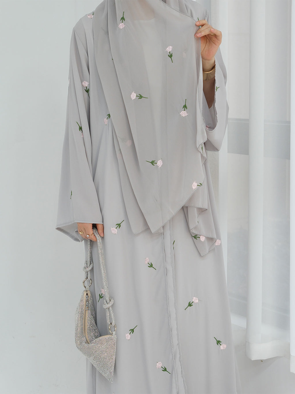 A person wearing a light gray modest outfit with a matching draped outer layer, decorated with small pink and green floral embroidery. The person is holding a matching light gray handbag against a bright white indoor background.