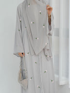 A person wearing a light gray modest outfit with a matching draped outer layer, decorated with small pink and green floral embroidery. The person is holding a matching light gray handbag against a bright white indoor background.
