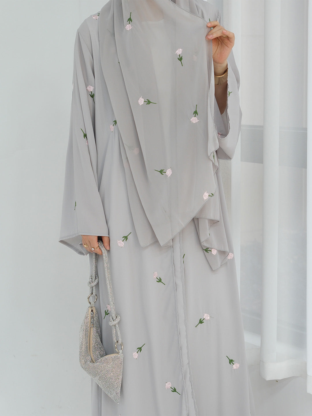 A person wearing a light gray modest outfit with a matching draped outer layer, decorated with small pink and green floral embroidery. The person is holding a matching light gray handbag against a bright white indoor background.