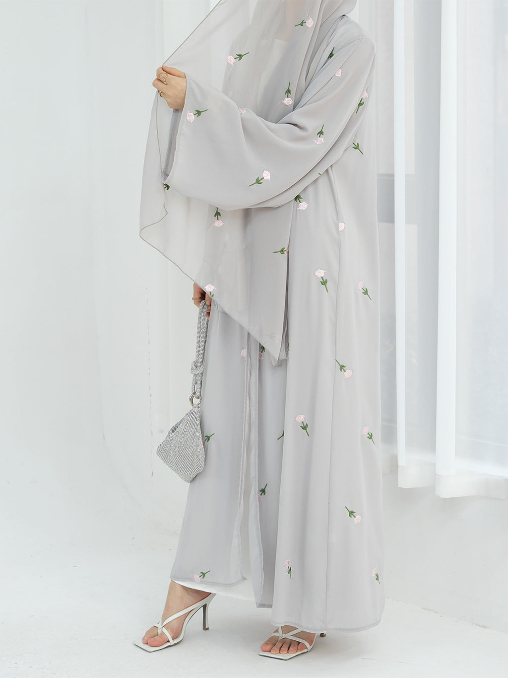 A woman wearing a light gray modest outfit with a long flowing abaya, matching wide-leg pants, a head covering, and white heeled sandals, holding a small gray handbag against a bright white indoor background.