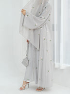 A woman wearing a light gray modest outfit with a long flowing abaya, matching wide-leg pants, a head covering, and white heeled sandals, holding a small gray handbag against a bright white indoor background.