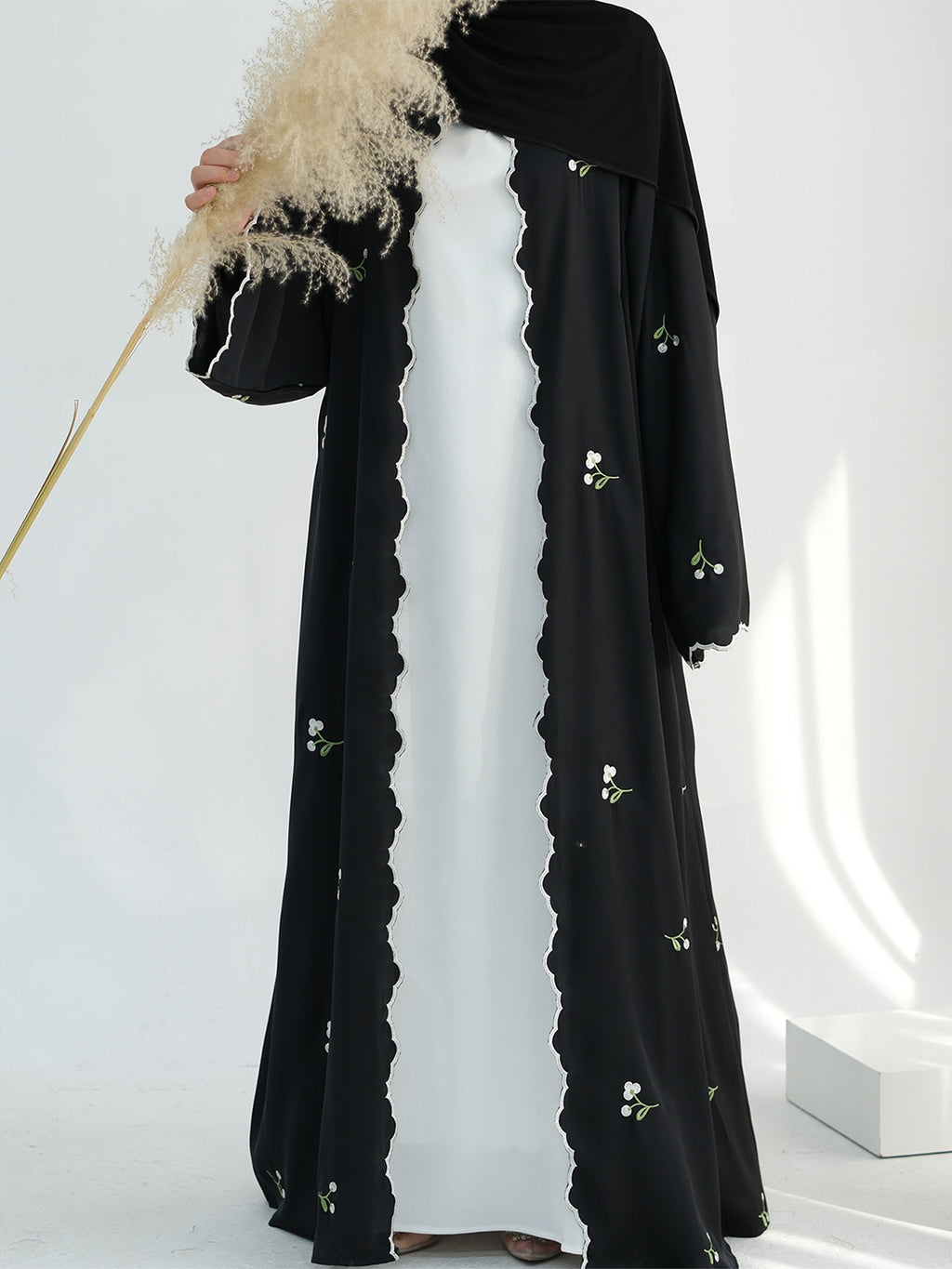 A woman wearing a black long abaya with scalloped edges and small floral embroidery, layered over a white dress, with a black headscarf. She is holding a decorative pampas grass stem in a bright white studio setting.