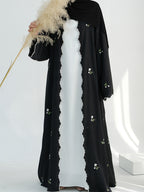 A woman wearing a black long abaya with scalloped edges and small floral embroidery, layered over a white dress, with a black headscarf. She is holding a decorative pampas grass stem in a bright white studio setting.