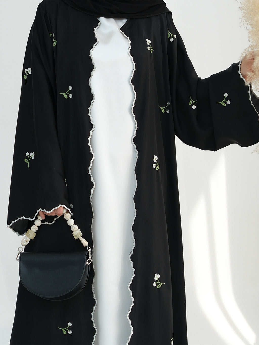A black open-front abaya or kaftan with embroidered white flowers and green stems, worn over a white dress. The garment has scalloped edges and is shown with a black handbag and pearl strap against a light studio background.
