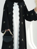 A black open-front abaya or kaftan with embroidered white flowers and green stems, worn over a white dress. The garment has scalloped edges and is shown with a black handbag and pearl strap against a light studio background.