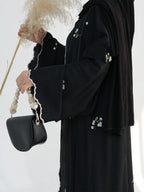 A person wearing a black abaya with white floral embroidery and scalloped white trim, holding a black handbag with a beaded strap and a dried pampas grass stem against a white studio background.