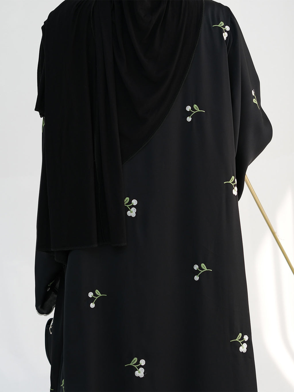 A black abaya dress with small white floral embroidery, shown on a mannequin with a matching draped black hijab against a plain white background.
