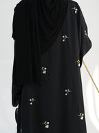A black abaya dress with small white floral embroidery, shown on a mannequin with a matching draped black hijab against a plain white background.