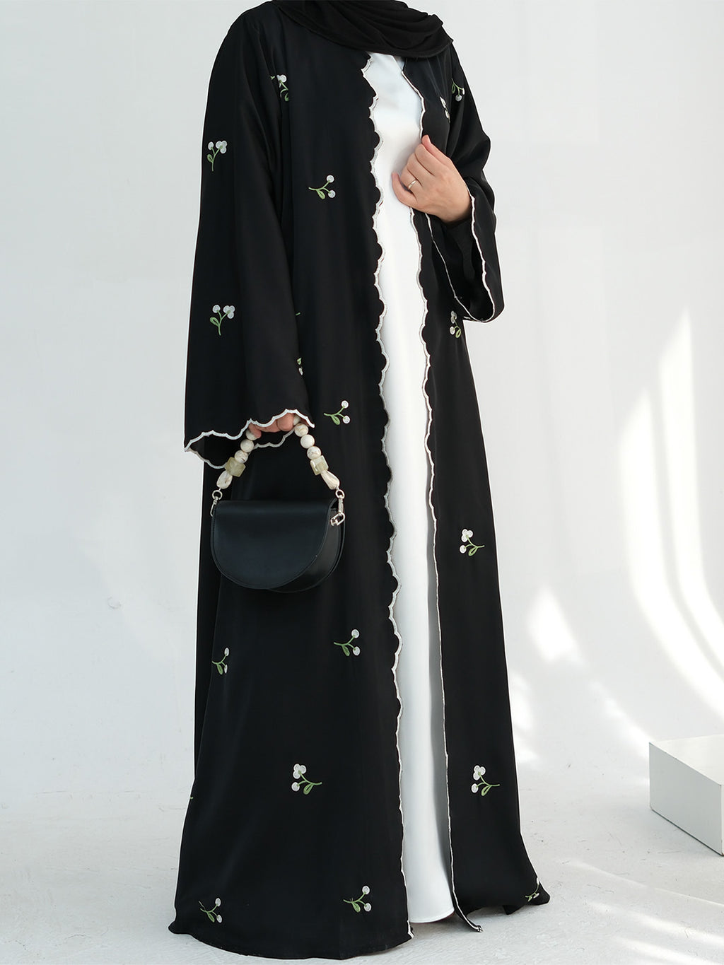 A woman models a long black abaya dress with delicate white floral embroidery, worn over a white inner dress and paired with a black headscarf and a small black handbag.