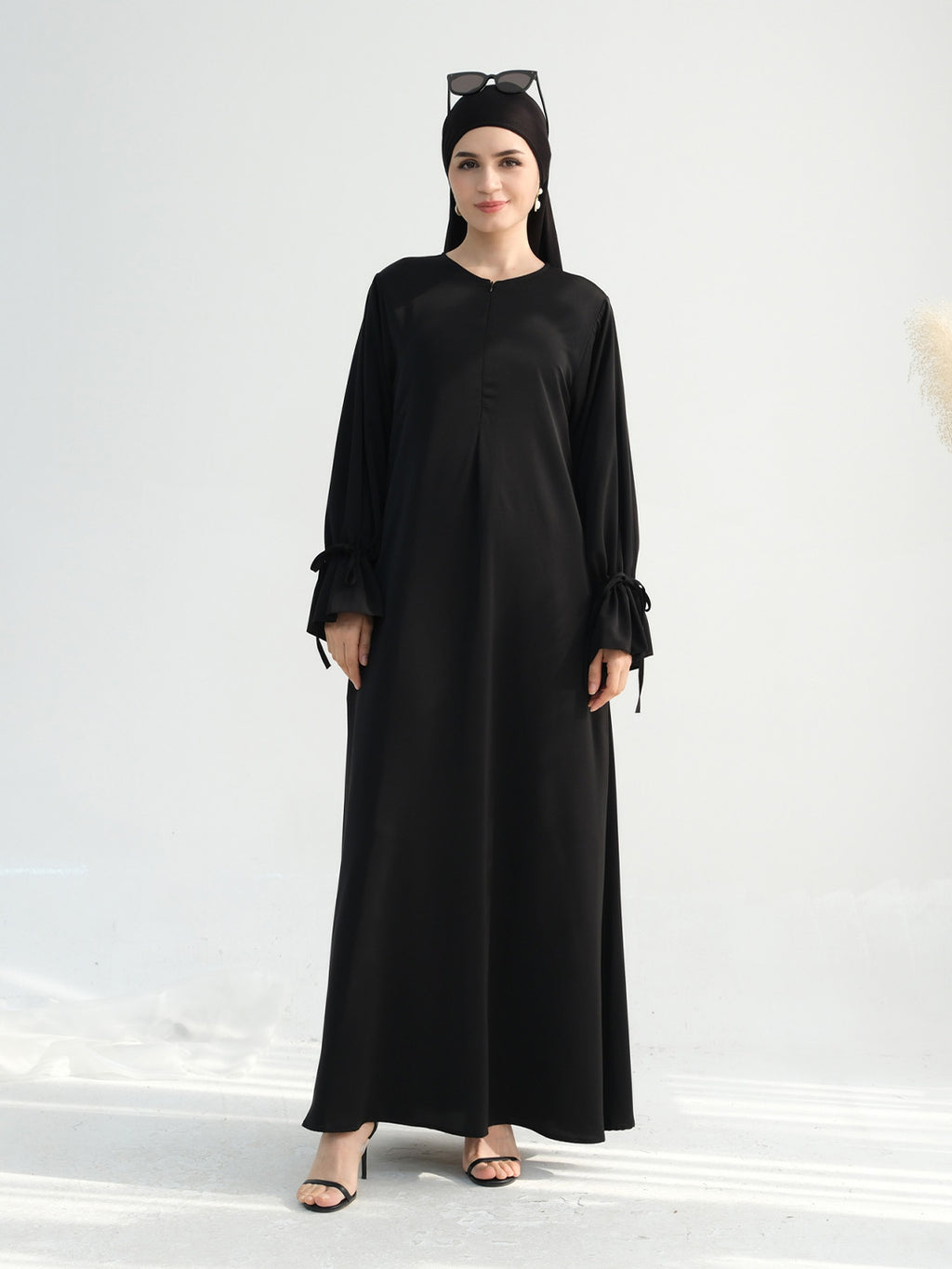 A woman modeling a long black abaya dress with long sleeves, tie details at the cuffs, and a round neckline, paired with a black headscarf and black sandals against a light gray studio background.