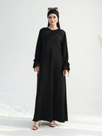 A woman modeling a long black abaya dress with long sleeves, tie details at the cuffs, and a round neckline, paired with a black headscarf and black sandals against a light gray studio background.