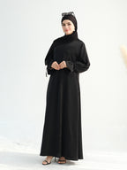 A woman modeling a long black modest dress with a high neck, long sleeves, and a matching black hijab, standing against a clean white studio background.