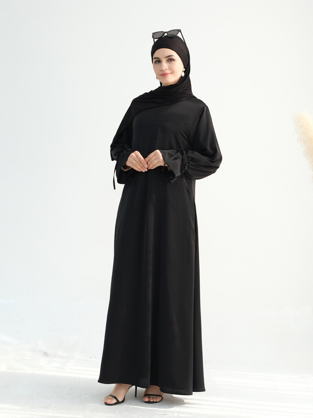 A woman modeling a long black modest dress with a high neck, long sleeves, and a matching black hijab, standing against a clean white studio background.