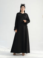 A woman modeling a long black abaya dress with a round neckline, long sleeves, and a flowing skirt, paired with a black headscarf and black sandals against a plain light background.