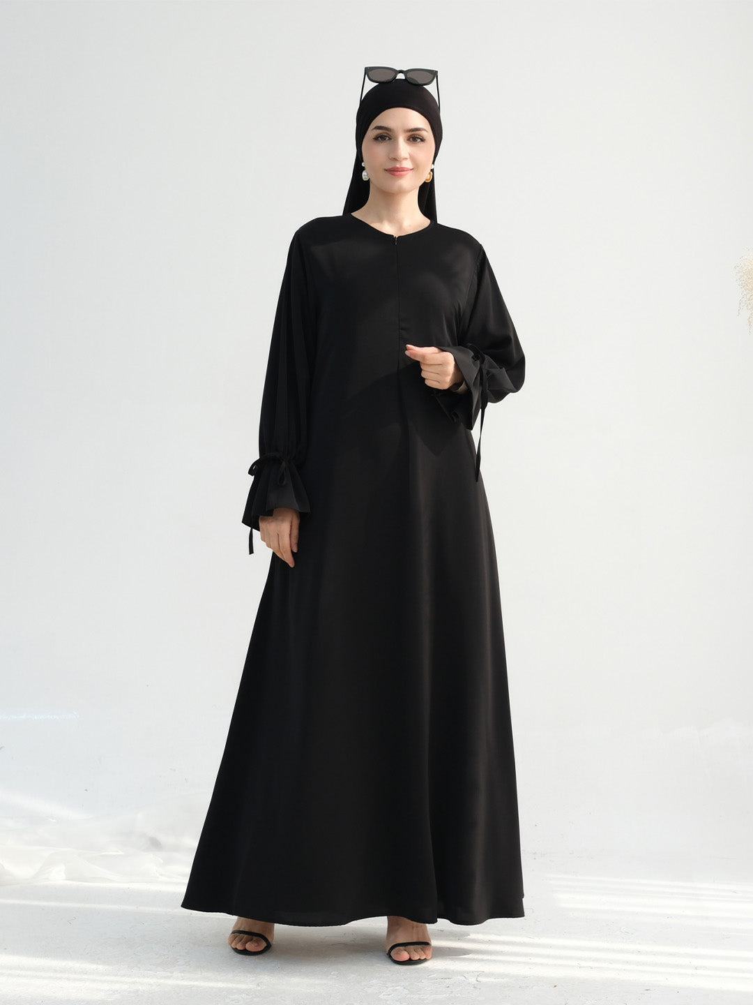 A woman modeling a long black abaya dress with a round neckline, long sleeves, and a flowing skirt, paired with a black headscarf and black sandals against a plain light background.