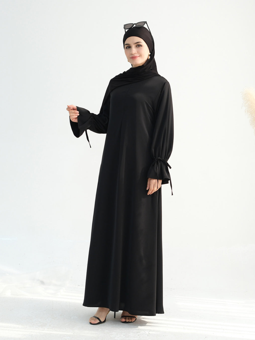 A woman modeling a long black abaya dress with wide sleeves and side ties, paired with a matching black hijab and black sandals, against a clean white studio background.