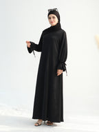 A woman modeling a long black abaya dress with wide sleeves and side ties, paired with a matching black hijab and black sandals, against a clean white studio background.