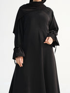 A black long-sleeved abaya dress with a draped shawl collar, tie details at the cuffs, and a matching black hijab, shown on a model against a plain white background.