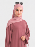 A woman wearing a dusty rose abaya and matching hijab, with black sunglasses resting on top of her head, photographed against a solid light gray background.