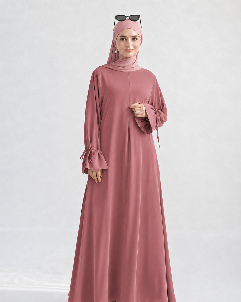 A woman wearing a dusty rose pink modest outfit, including a long flowing abaya dress with flared sleeves, a matching hijab, and sunglasses resting on her head, photographed against a light gray studio background.