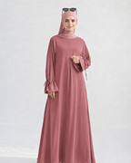 A woman wearing a dusty rose pink modest outfit, including a long flowing abaya dress with flared sleeves, a matching hijab, and sunglasses resting on her head, photographed against a light gray studio background.