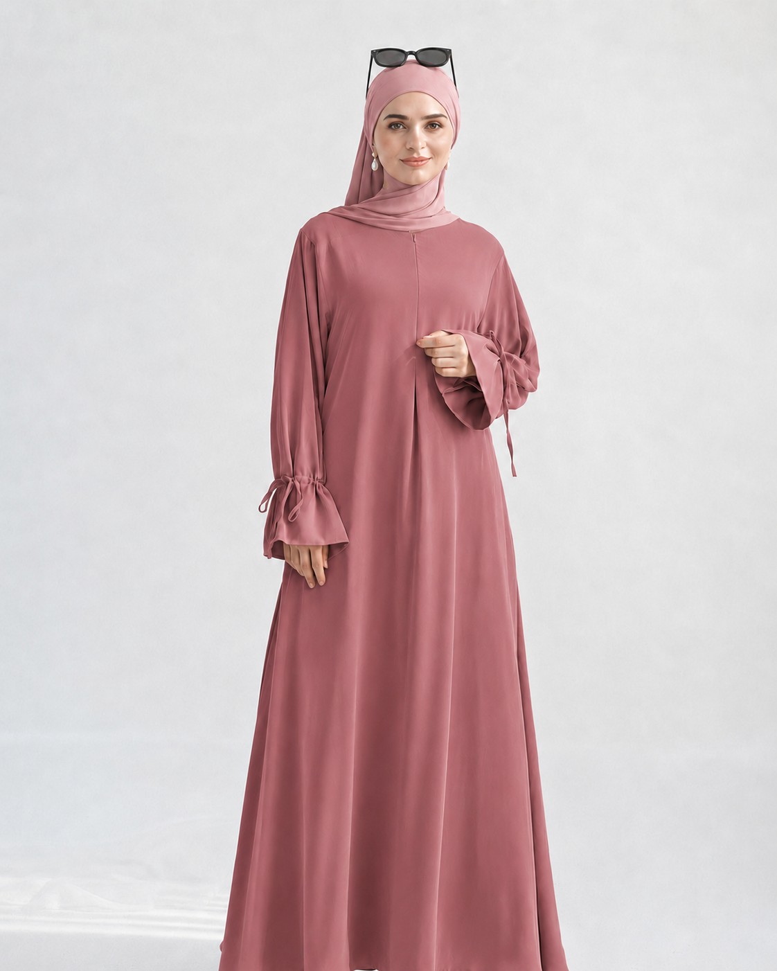A woman wearing a dusty rose pink modest outfit, including a long flowing abaya dress with flared sleeves, a matching hijab, and sunglasses resting on her head, photographed against a light gray studio background.