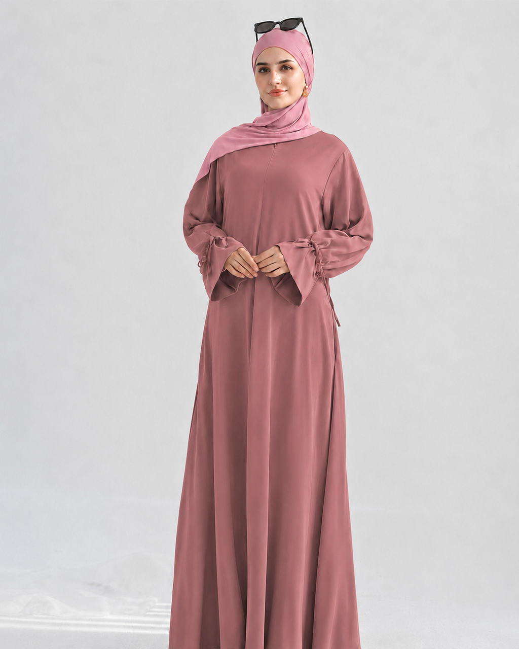 A woman modeling a long-sleeved dusty rose abaya with matching hijab and sunglasses on her head, set against a plain light gray studio background.