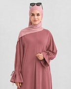 A woman wearing a dusty rose abaya with matching hijab and sunglasses on her head, posing against a light gray studio background.