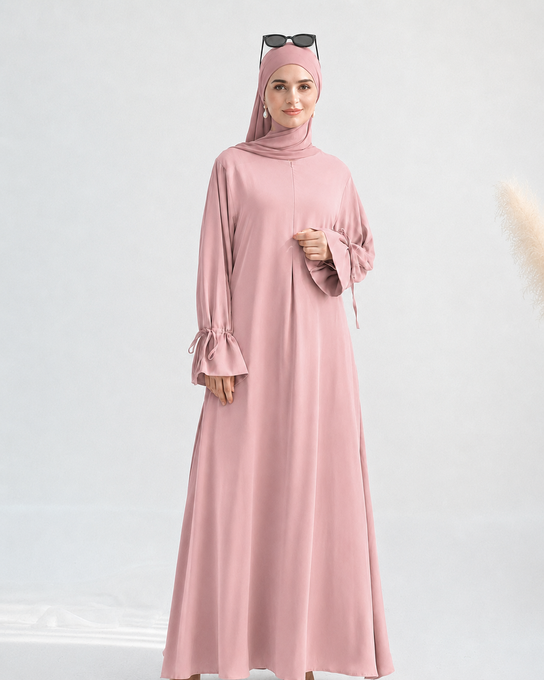 A woman modeling a dusty pink long-sleeve maxi abaya with a matching hijab and sunglasses on her head, set against a light gray studio background.