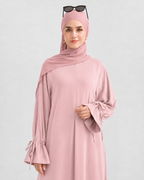 A woman wearing a dusty pink modest outfit consisting of a long-sleeved abaya with tie details on the cuffs, a matching hijab, and sunglasses resting on her head, set against a plain light gray background.