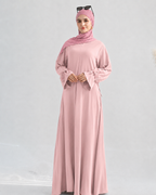 A woman modeling a long-sleeved dusty pink abaya with matching hijab and sunglasses on her head, set against a plain light gray background.