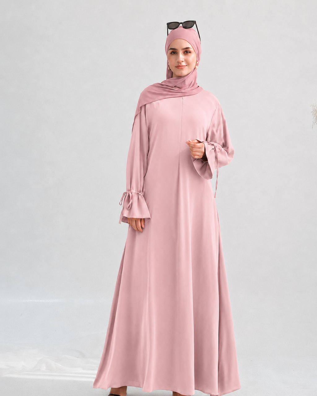 A woman modeling a long-sleeved dusty pink abaya dress with a matching hijab and sunglasses on her head, standing against a light gray studio background.