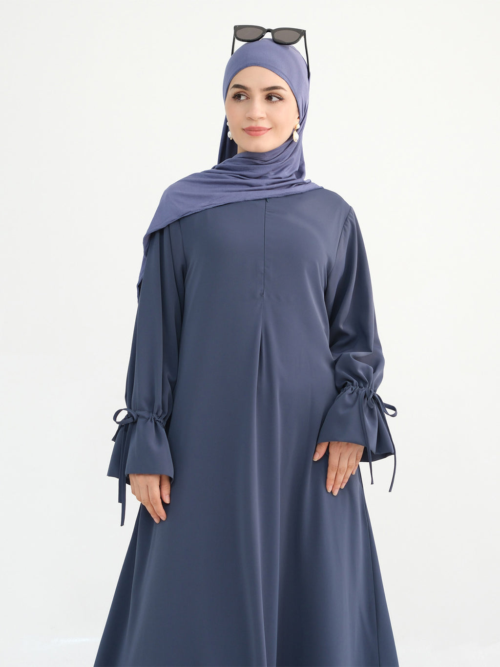 A woman modeling a slate blue long-sleeve abaya with tie details on the cuffs, paired with a matching hijab and sunglasses resting on top of her head, against a plain white background.