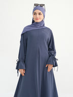 A woman modeling a slate blue long-sleeve abaya with tie details on the cuffs, paired with a matching hijab and sunglasses resting on top of her head, against a plain white background.