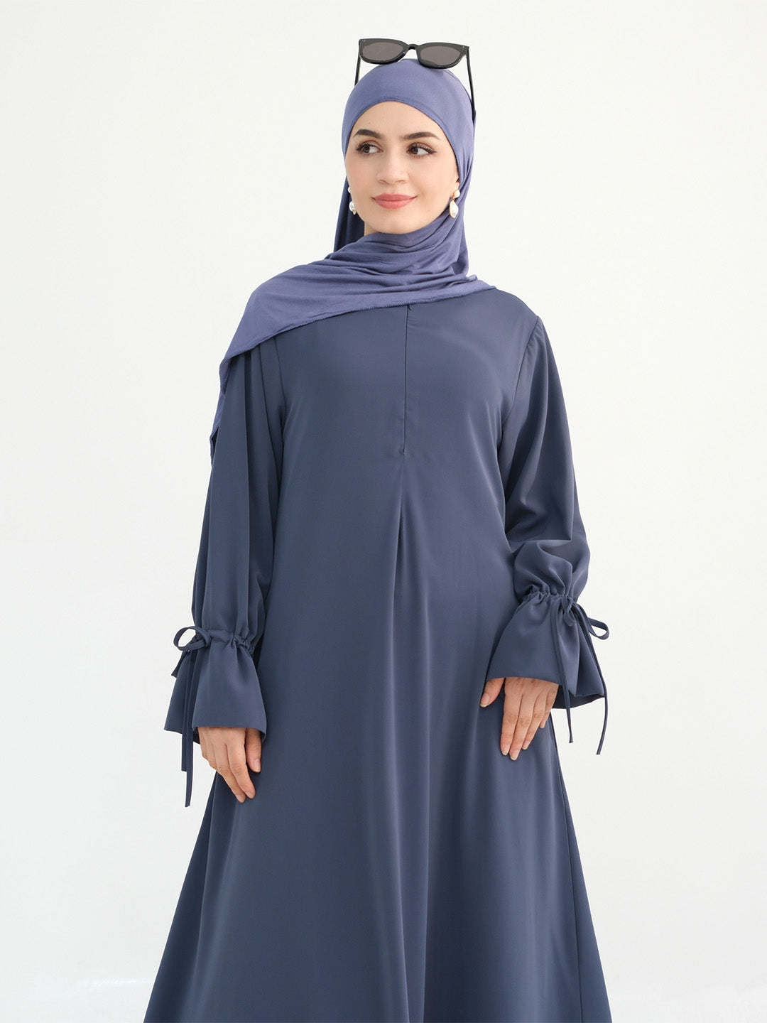 A woman modeling a slate blue long-sleeve abaya with tie details on the cuffs, paired with a matching hijab and sunglasses resting on top of her head, against a plain white background.