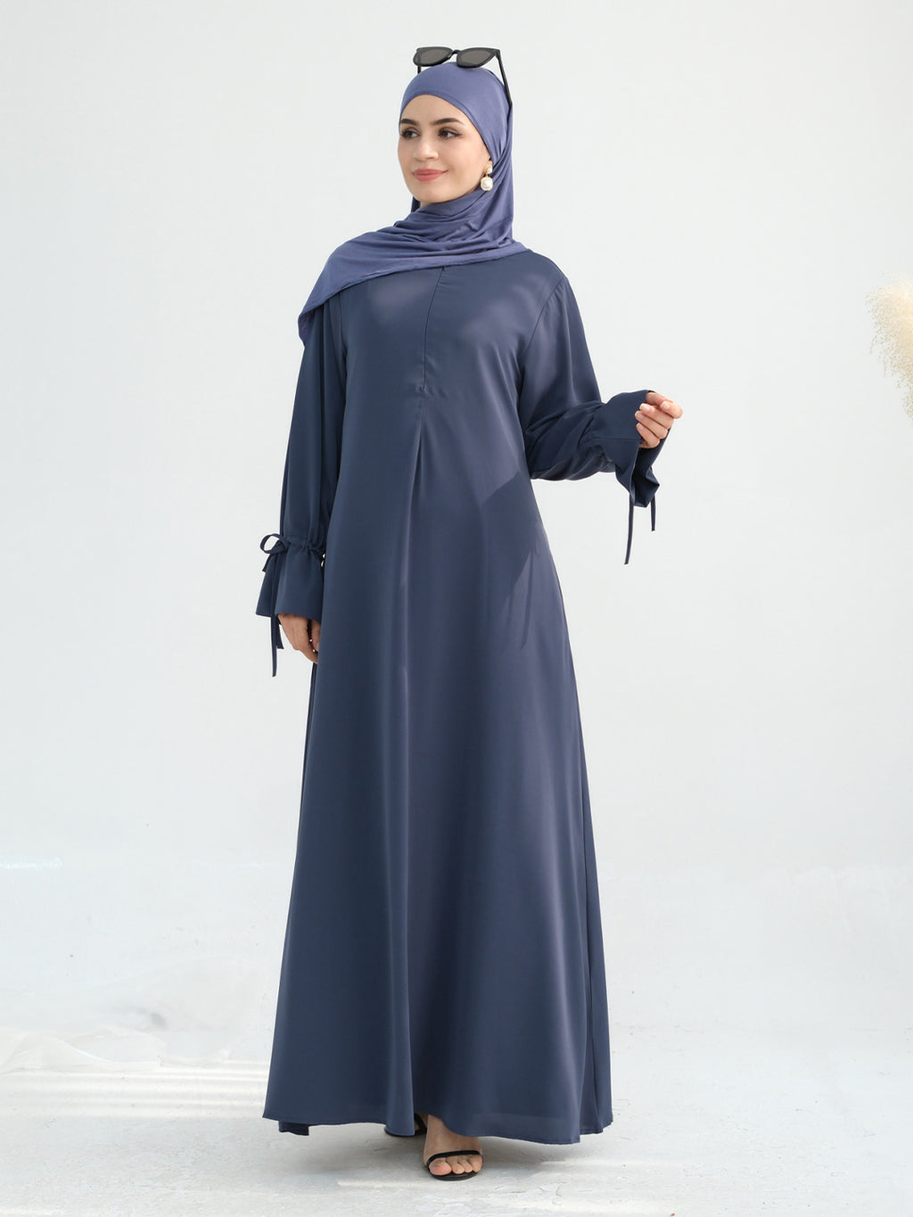 A woman modeling a long-sleeved navy blue abaya dress with side tie details, paired with a matching navy blue hijab and black sandals, set against a plain light gray background.