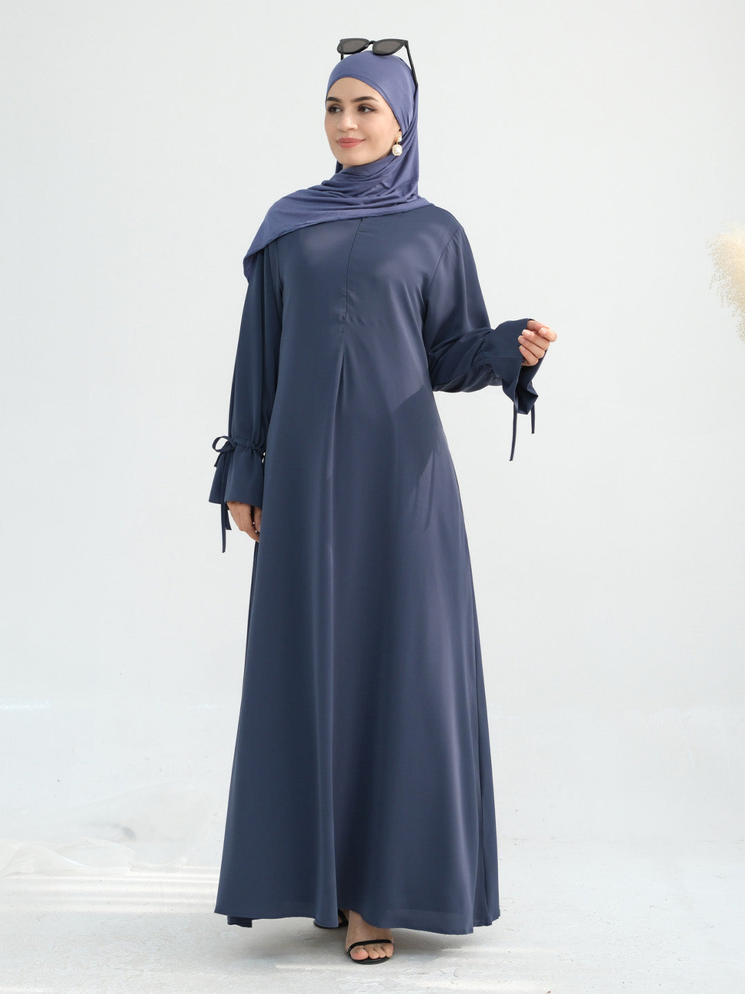 A woman modeling a long-sleeved navy blue abaya dress with side tie details, paired with a matching navy blue hijab and black sandals, set against a plain light gray background.