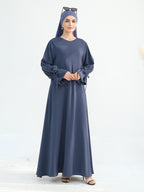 A woman modeling a long navy blue abaya dress with a matching headscarf and sunglasses on top, standing against a plain white studio background.