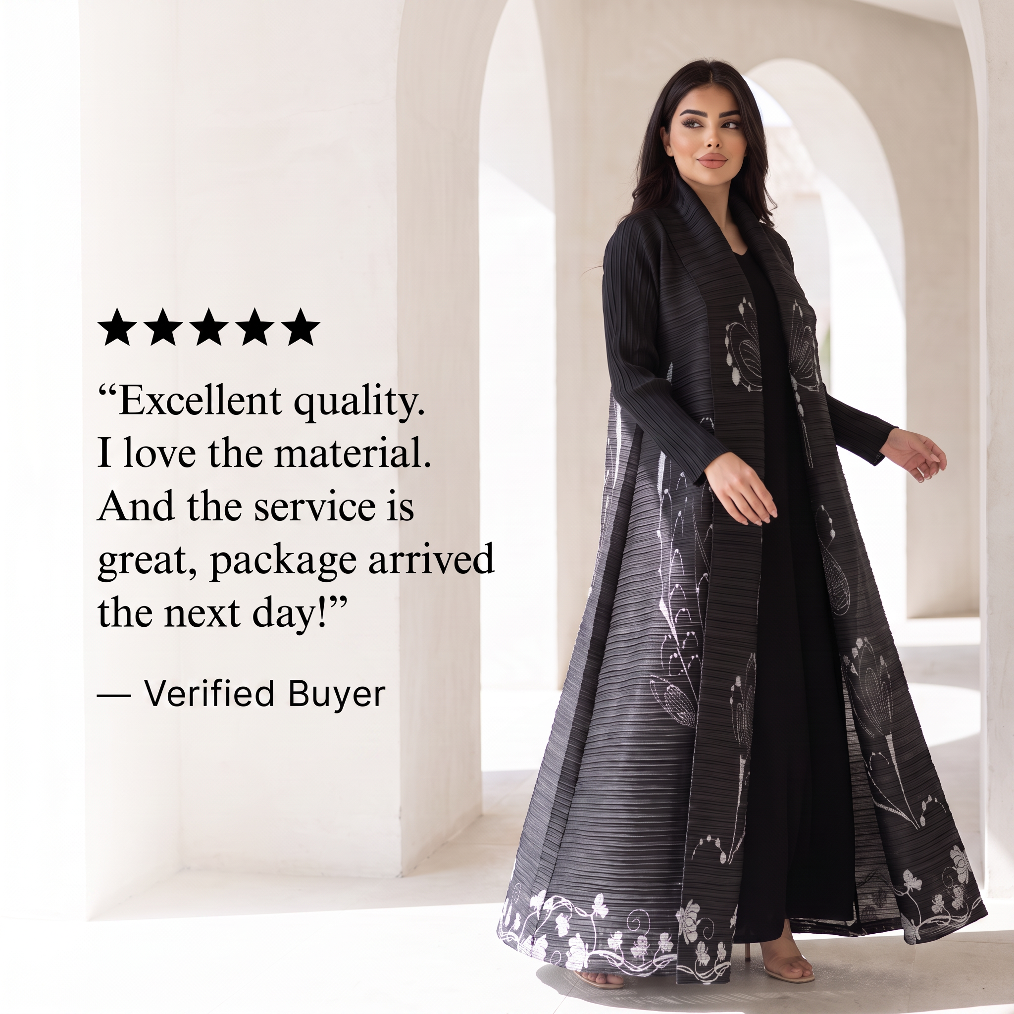 A woman models a long black outer garment with gray striped and white floral patterns over a black outfit, standing in a bright white arched corridor. A five-star review quote is printed on the left side of the image.