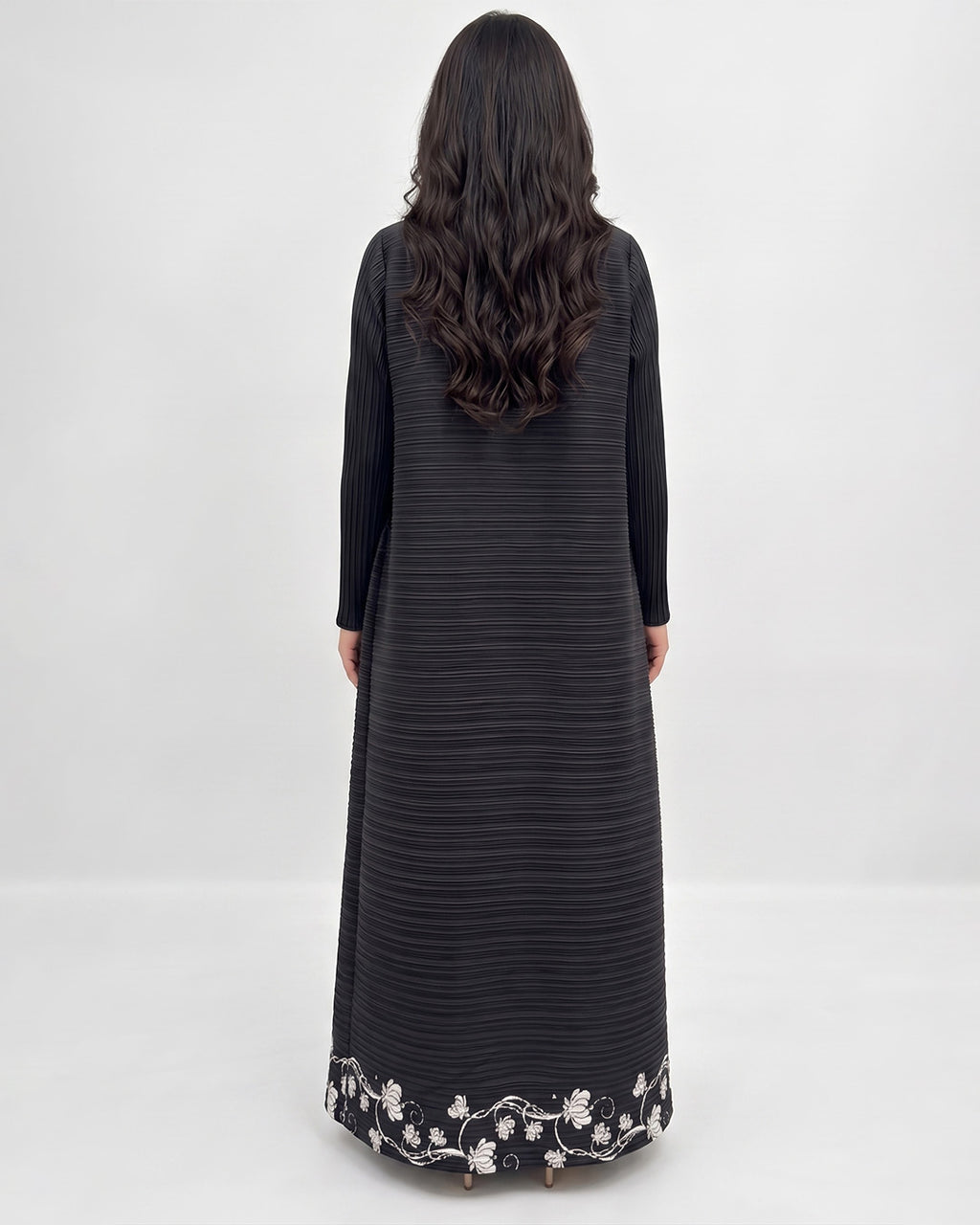A woman viewed from behind wearing a long black ribbed dress with long sleeves and a white floral embroidered border at the hem, against a plain light gray studio background.